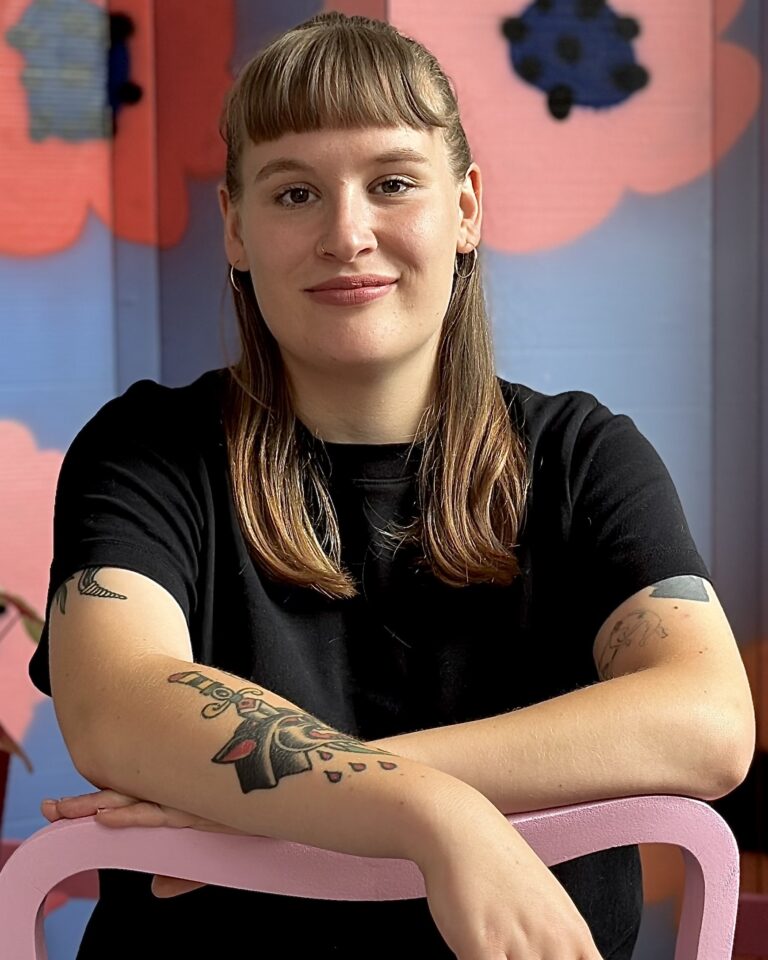 Eine Person sitzt auf einem Stuhl vor einer bunten Wand mit großen Blumenmotiven.