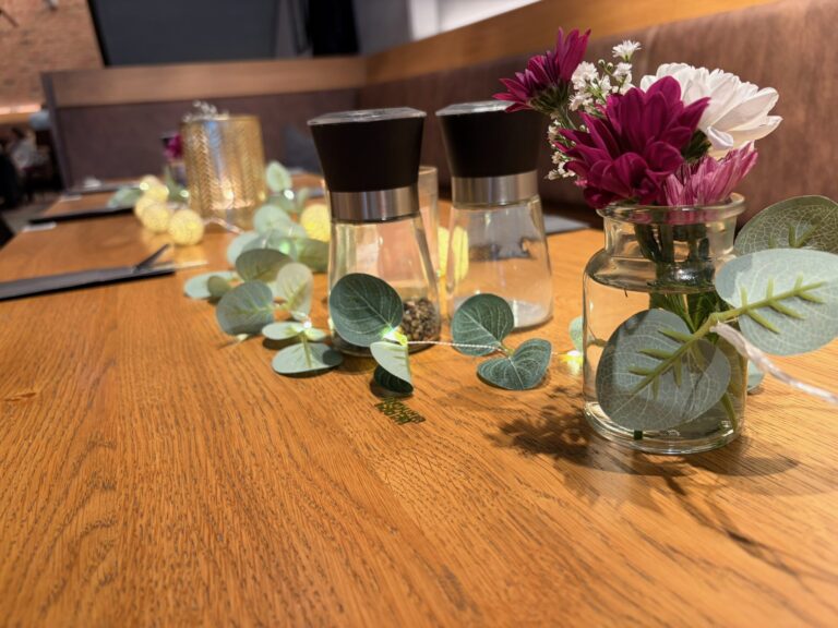 Detailaufnahme eines dekorierten Tisches im Café Anders mit Gewürzmühlen, Blumen in Vasen und dekorativer Lichterkette.
