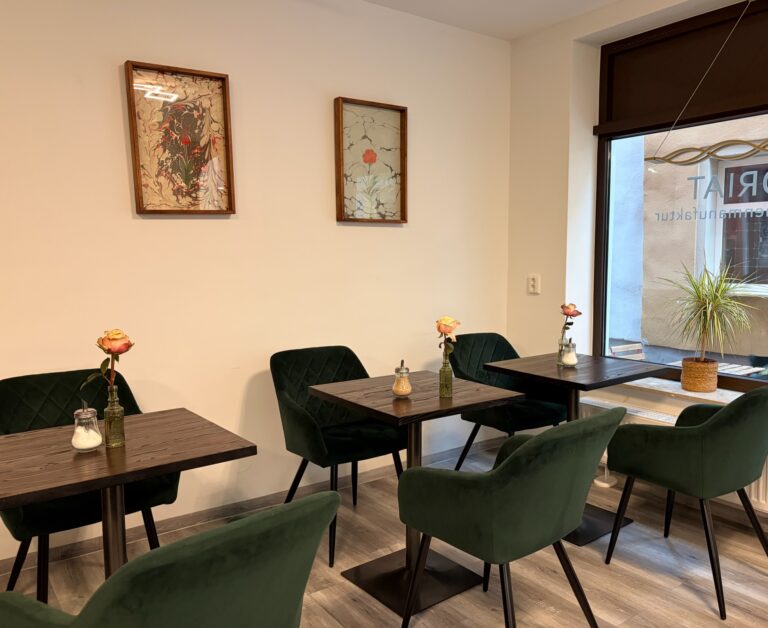 Stilvolle Sitzecke in der Koriat Kuchenmanufaktur mit dunklen Tischen, samtigen, grünen Stühlen und Blumen.