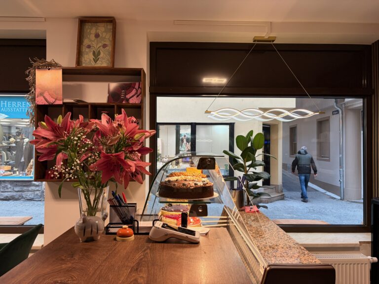 Innenansicht der Koriat Theke mit Kuchenauslage und Blumen, Blick durch das große Fenster auf die Straße.