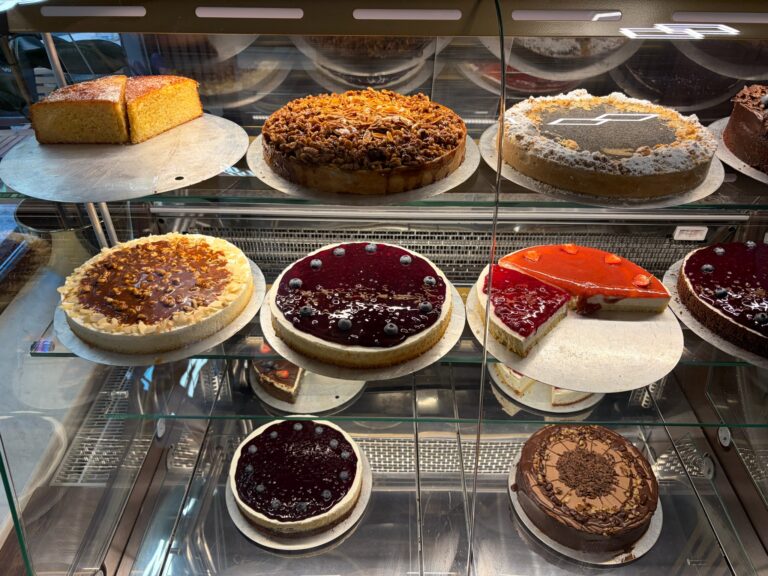 Nahaufnahme der Kuchenvitrine von Koriat mit verschiedenen Kuchen, darunter Beeren-Cheesecake, Mandel-Streuselkuchen und Erdbeertarte.