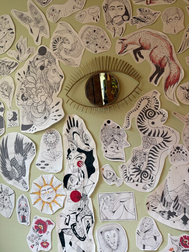 Eine Studiowand voller handgezeichneter Tattoo-Motive und Illustrationen in Schwarz-Weiß und Farbe. In der Mitte hängt ein dekorativer Spiegel in Form eines Auges.