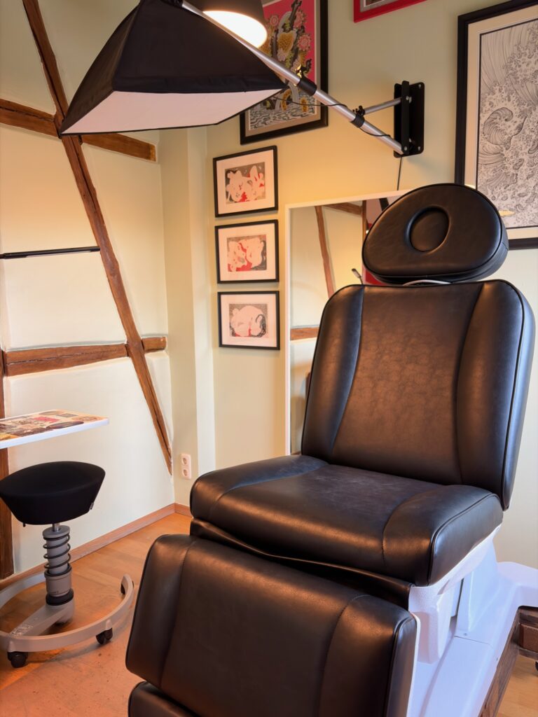 Heller, freundlicher Arbeitsbereich in einem Tattoostudio mit einem weißen Behandlungsstuhl, warmem Licht und zahlreichen Kunstzeichnungen an der Wand.