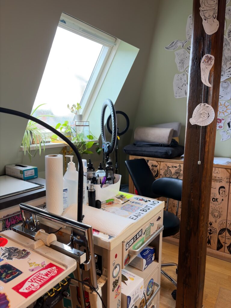 Ein heller, gut organisierter Arbeitsplatz in einem Tattoo- oder Designstudio, mit Arbeitsutensilien wie Handschuhe, Desinfektionsmittel, Küchenpapier, Tätowierfarben und künstlerischen Zeichnungen im Hintergrund.