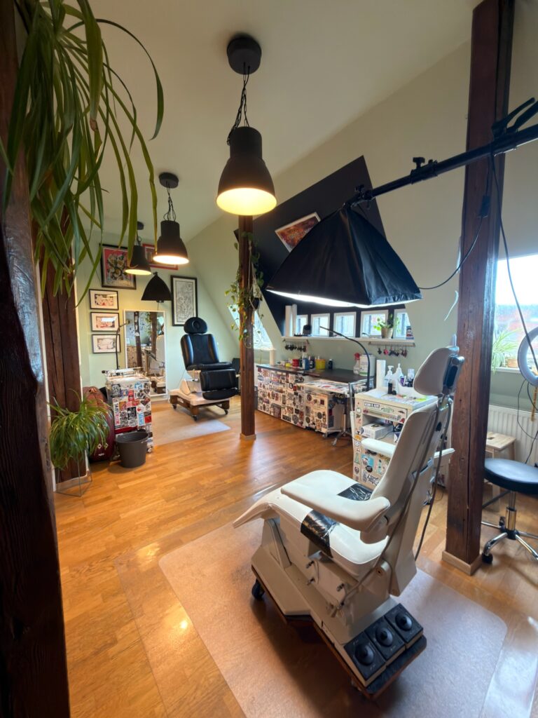 Ein heller Tattoo-Arbeitsraum mit zwei Behandlungsliegen, warmem Holzfußboden, individuellen Dekorationen und Arbeitsmaterialien, eingerichtet in einem gemütlichen Studio mit Dachschrägen und Pflanzen.