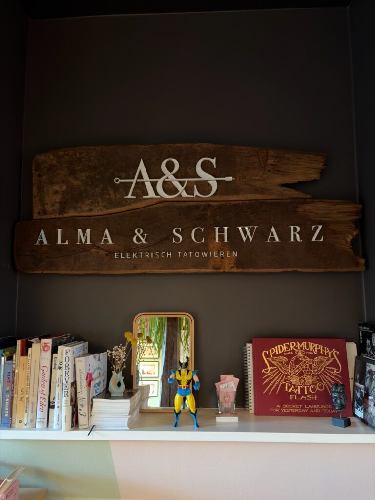 Ein dekoratives Wanddisplay mit dem Studio-Logo „Alma & Schwarz – Elektrisch Tätowieren“ auf einer rustikalen Holztafel, darunter ein Regal mit Büchern, einem Spiegel und kleinen Deko-Objekten.