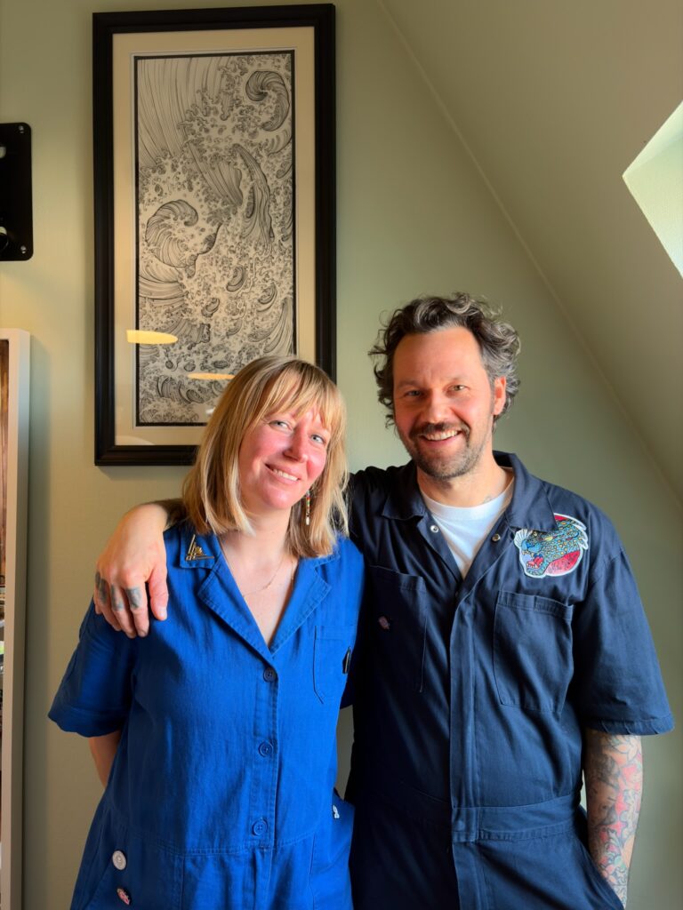 Zwei Personen (Sandy und Andreas) stehen nebeneinander in ihrem Tattoostudio, beide lächeln freundlich. Im Hintergrund hängt ein großformatiges schwarz-weißes Kunstwerk mit Wellenmotiv.