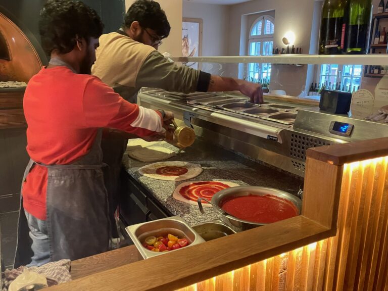 Zwei Pizzabäcker stehen hinter der Theke und verteilen konzentriert Tomatensauce auf dem Pizzateig.