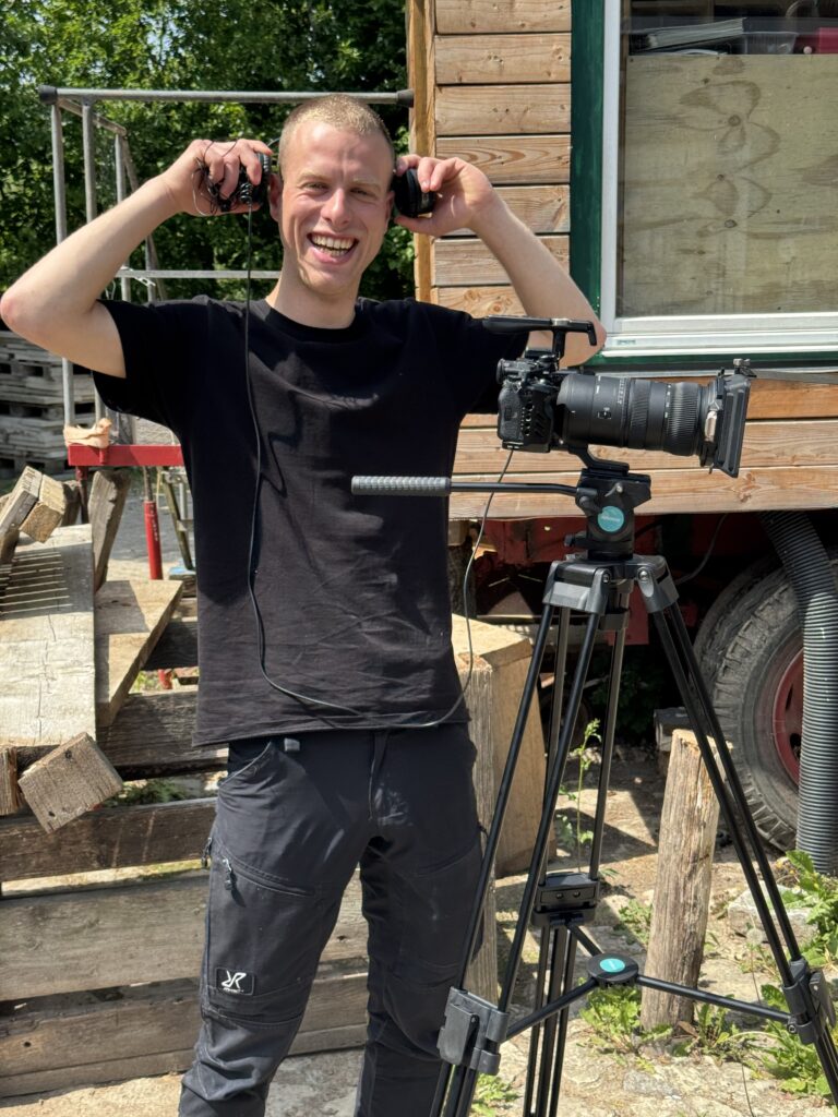 Ein junger Videograf steht lachend neben einer professionellen Kamera auf einem Stativ im Freien und hält sich die Kopfhörer an die Ohren; im Hintergrund ein hölzerner Bauwagen.
