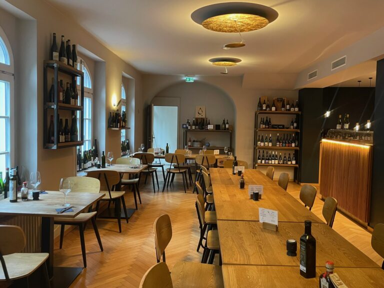 Blick in den stilvollen Gastraum mit Fischgrät-Parkett, hellen Holztischen und modernen, goldenen Deckenleuchten.