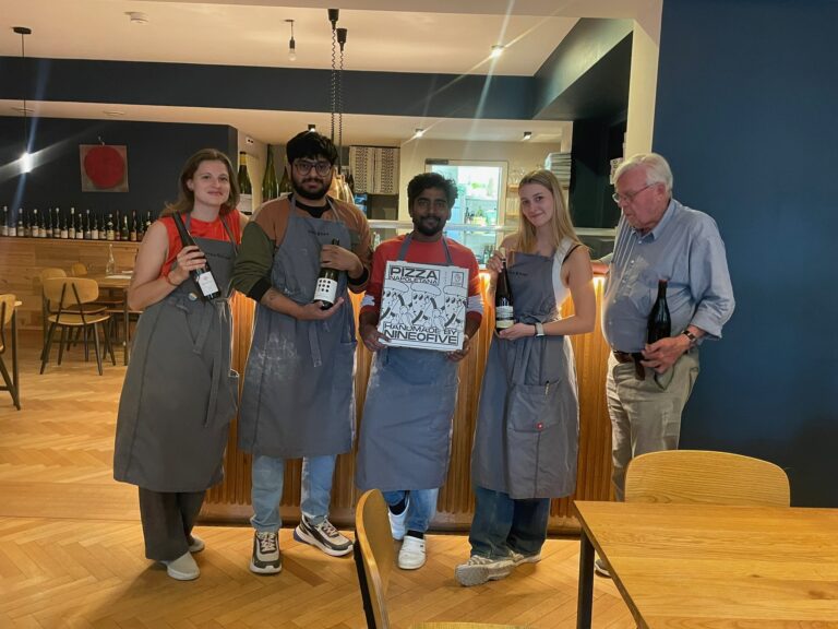 Das Team von Nine O'five steht lächelnd zusammen, hält Weinflaschen und einen Pizzakarton mit dem Logo in die Kamera.