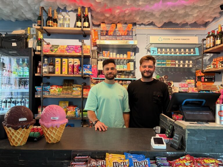 Inhaber der Trinkstube stehen freundlich hinter dem Tresen, umgeben von Regalen mit Snacks, Spirituosen und einer dekorativen Wolkendecke.