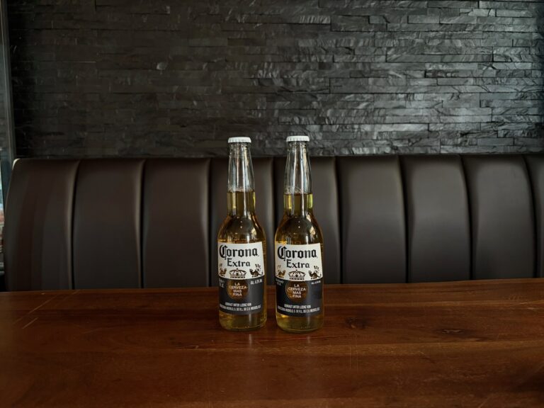 Zwei Corona-Bierflaschen stehen auf einem massiven Holztisch vor einer dunklen Steinwand in der Trinkstube Jena.