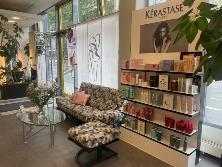 Gemütlicher Wartebereich mit gemustertem Sofa und Glas-Tisch neben einem Regal mit Kérastase-Pflegeprodukten bei Friseur CC.