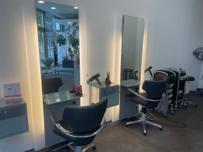 Moderne Friseur-Bedienplätze mit großen, beleuchteten Spiegeln und bequemen schwarzen Sesseln im Salon Friseur CC.