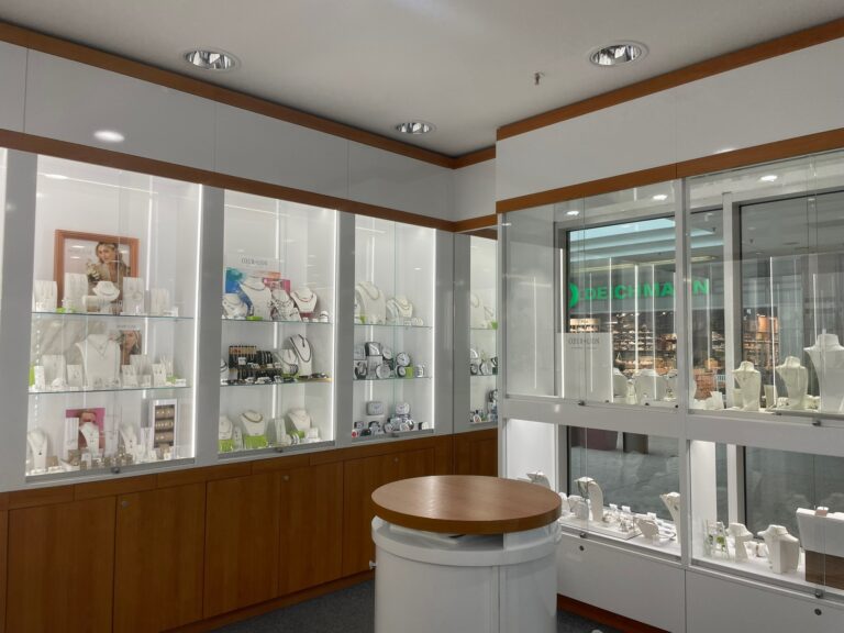 Innenansicht des hell erleuchteten Schmuckgeschäfts mit weißen Glasvitrinen entlang der Wände und Blick in die Einkaufspassage.