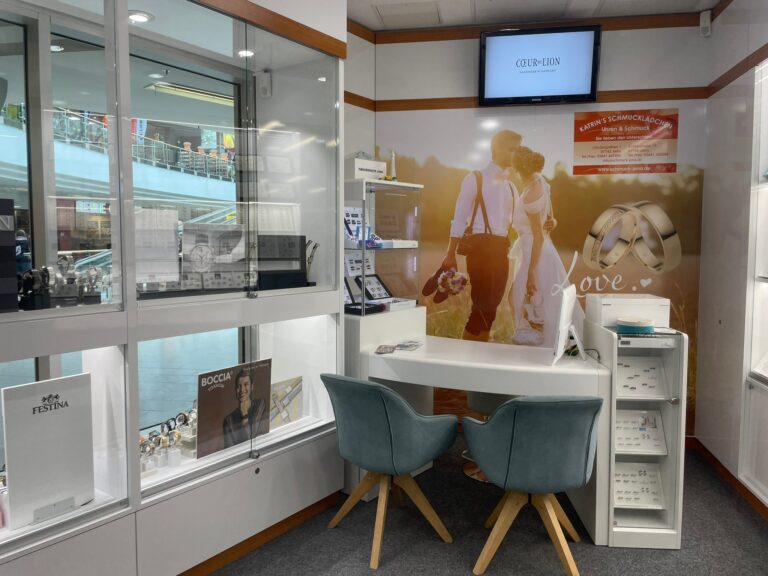 Gemütliche Beratungsecke für Trauringe mit zwei blauen Samtsesseln, einem weißen Tisch und einem großen Wandbild eines Brautpaares.