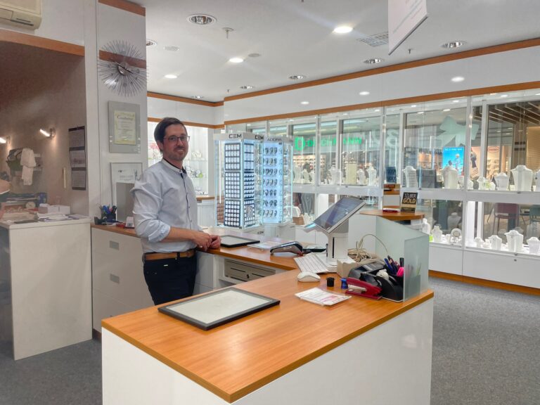 Mitarbeiter steht lächelnd im Verkaufsraum neben gerahmten Urkunden und einem Gesellenbrief, die an der Wand hängen.