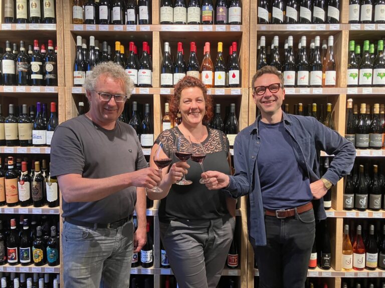 Das dreiköpfige Team von Biowein-erlesen steht lachend vor einem gut gefüllten Weinregal und stößt mit Rotweingläsern an.
