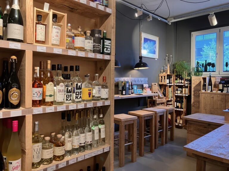 Gemütliche Ecke mit Holzhockern an einem Wandtresen unter blauer Kunst, im Fenster sind Silhouetten von Weinflaschen zu sehen.