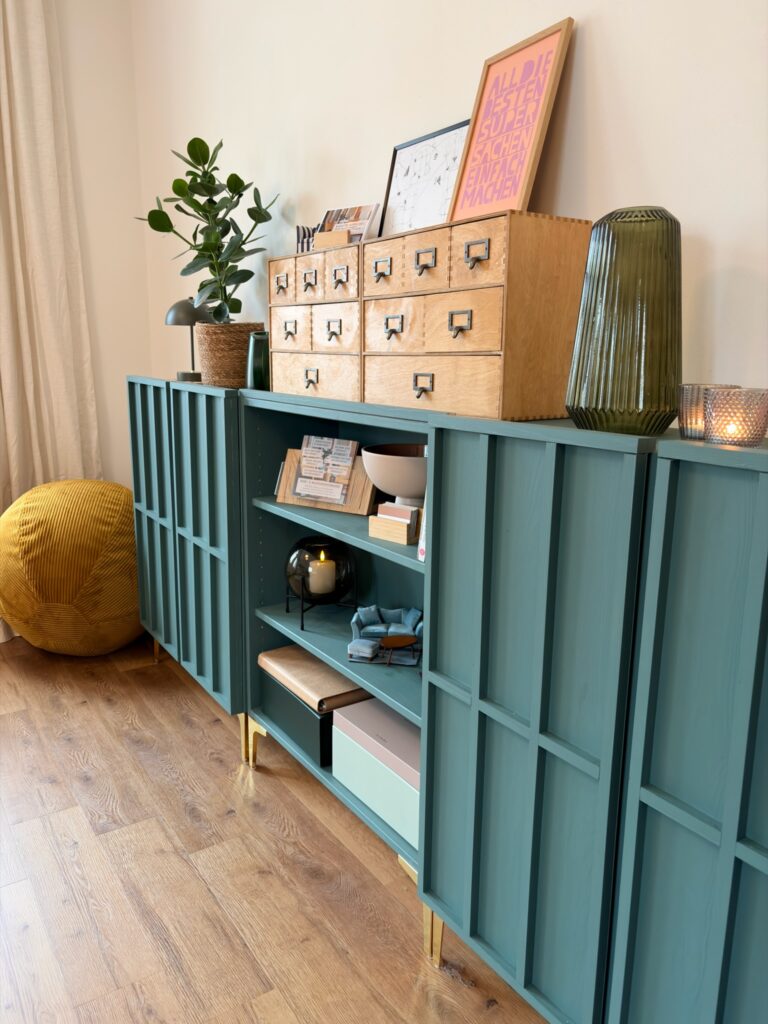 Ein petrolblaues Sideboard mit Lamellentüren, auf dem zwei hölzerne Schubladenelemente sowie eine grüne Glasvase und Pflanzen platziert sind.