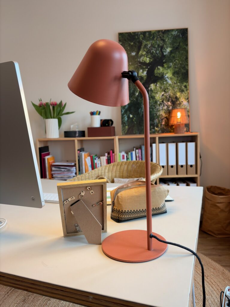 Ein weißer Schreibtisch im Vordergrund mit einer terracottafarbenen Lampe, einem Bilderrahmen und einem Etui. Im unscharfen Hintergrund ist ein Regal mit Aktenordnern erkennbar.