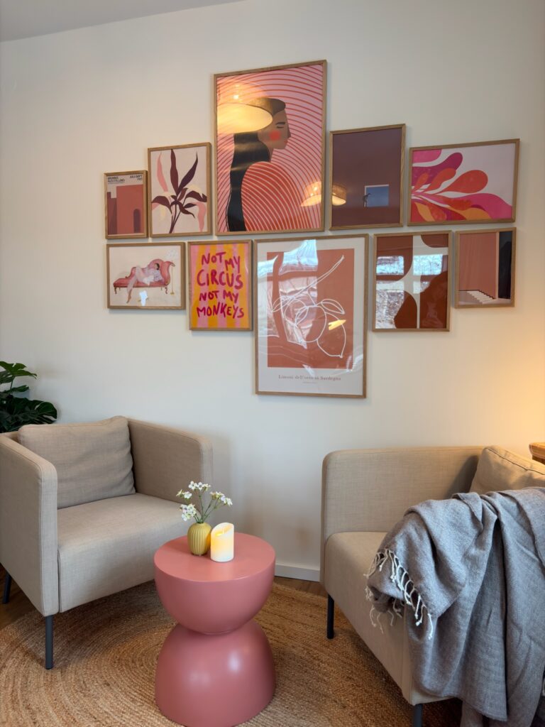 Zwei helle Polstersessel stehen sich an einem runden, rosa Beistelltisch gegenüber. An der Wand dahinter hängt eine symmetrisch angeordnete Galerie aus verschiedenen Kunstdrucken in Holzeinfassungen.