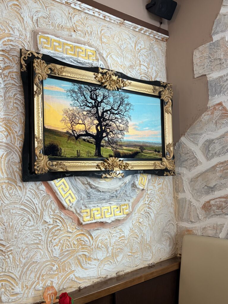 Ein goldgerahmtes Landschaftsbild hängt an einer dekorativen Wand mit Mustern. Das Gemälde zeigt einen kahlen schwarzen Baum, der auf einer Wiese steht. Der Himmel ist blau und gold verfärbt und stellt einen Sonnenaufgang dar.