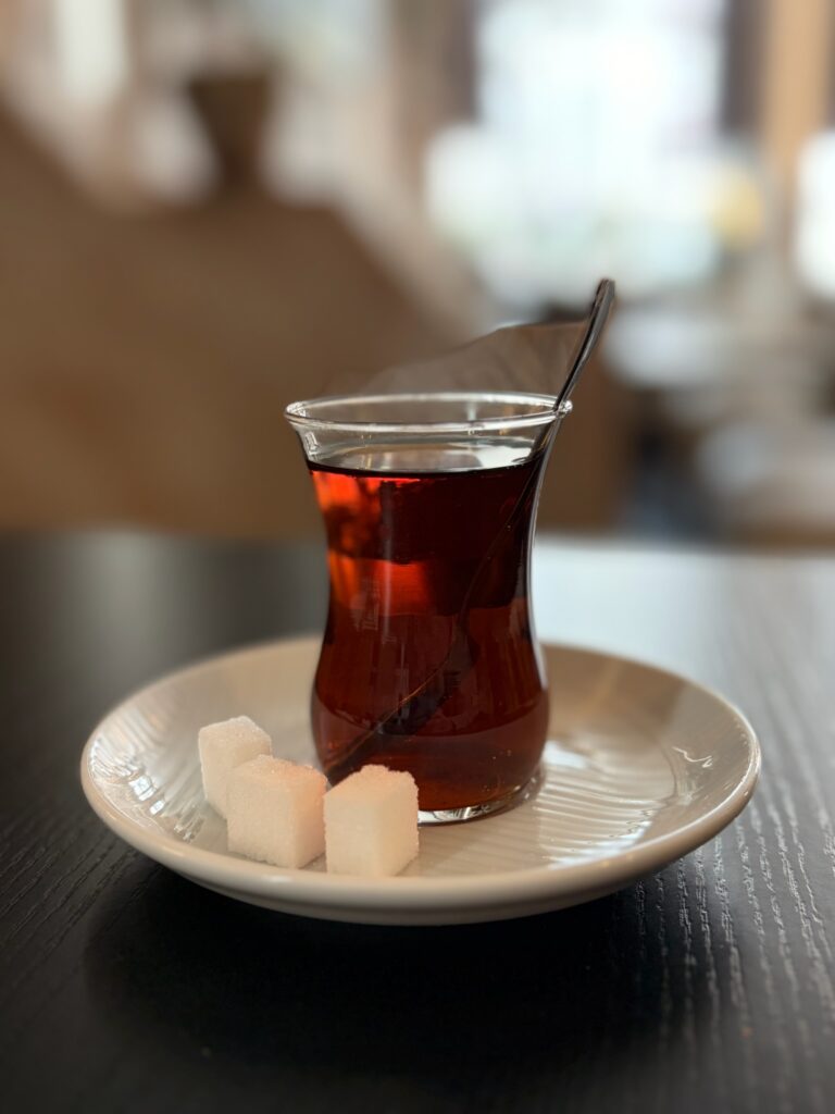 Glas mit schwarzem Tee und Löffel steht auf einer Untertasse mit Zuckerwürfeln auf einem schwarzen Tisch.