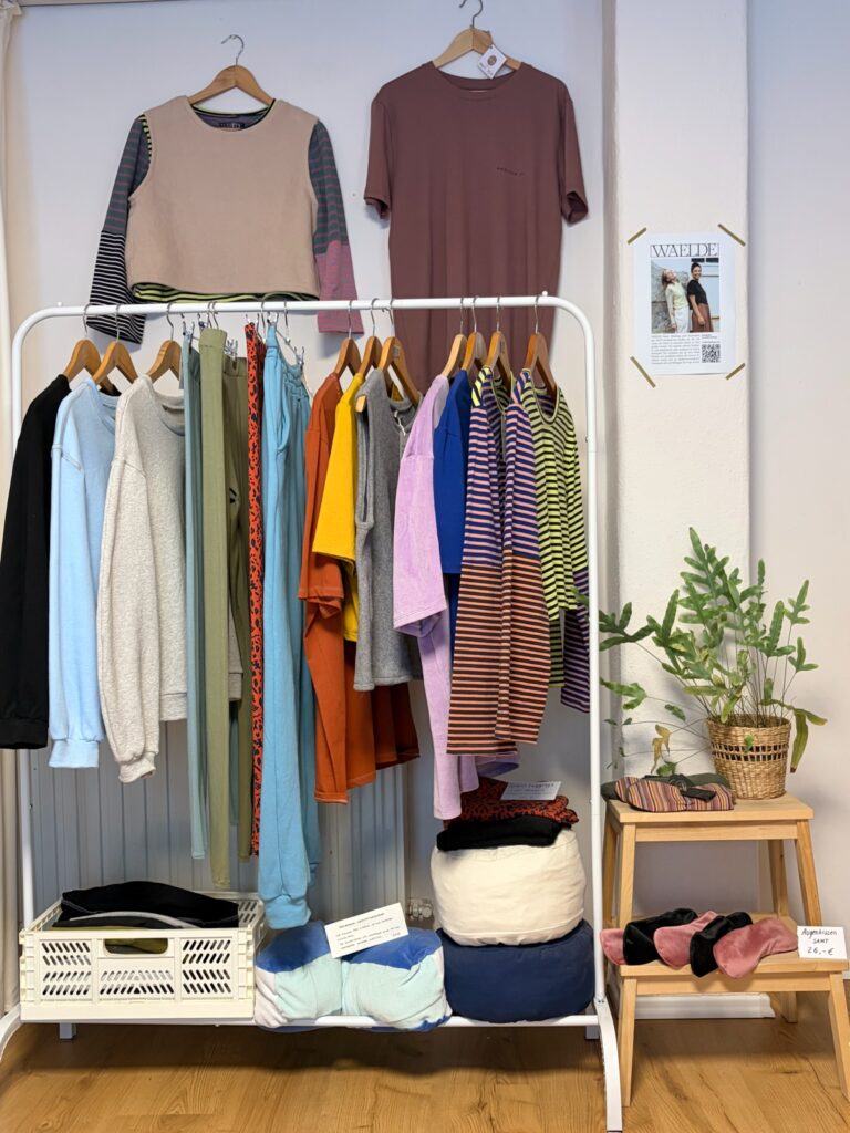 Eine Kleiderstange mit bunter, nachhaltiger Kleidung des Labels Waelde neben Yogakissen und handgemachten Ohrringen.
