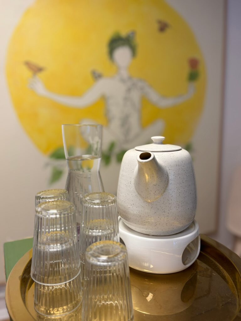 Eine weiße Teekanne auf einem Stövchen neben Gläsern auf einem goldenen Tablett, im Hintergrund ein gelbes Wandgemälde einer meditativen Figur.
