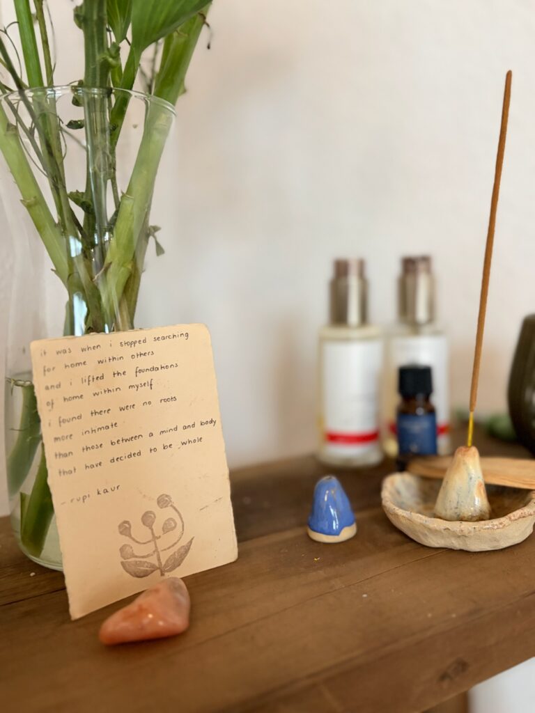 Stillleben auf einem Holzregal mit einer Postkarte von Rupi Kaur, einer kleinen Meridian-Figur, ätherischen Ölen und einem brennenden Räucherstäbchen.