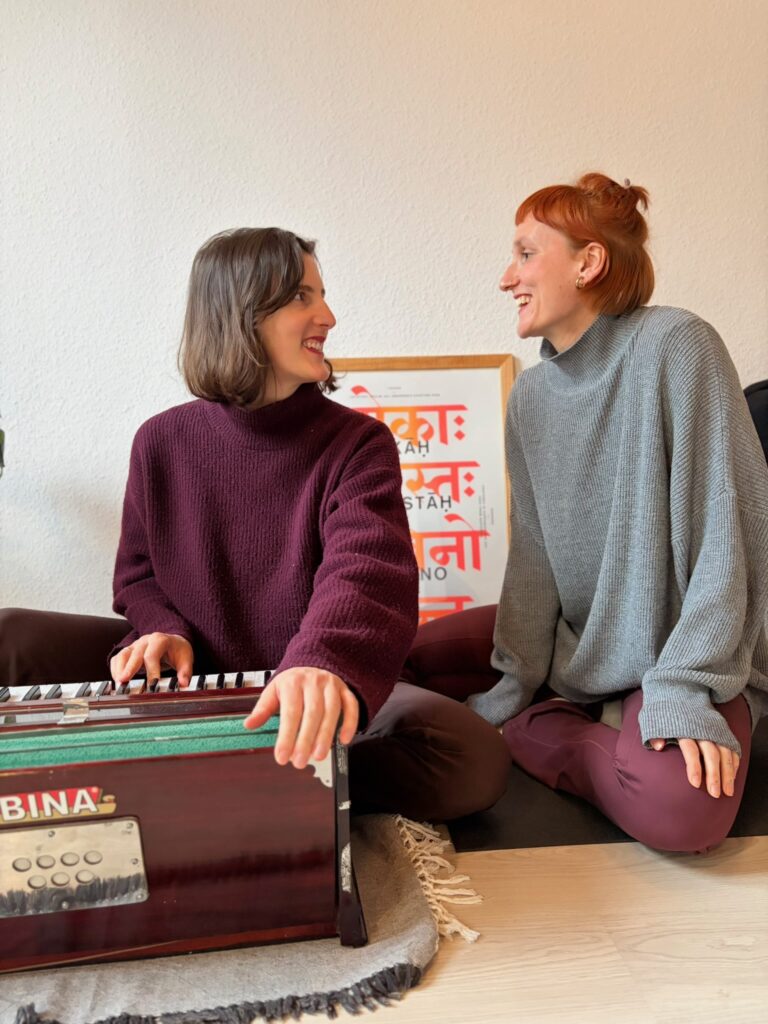 Zwei Frauen sitzen lächelnd am Boden, eine spielt ein hölzernes Harmonium der Marke Bina.