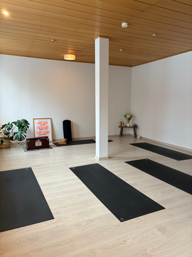 Heller Raum mit Holzdecke und hellem Boden, auf dem fünf schwarze Yoga-Matten ausgerichtet sind.