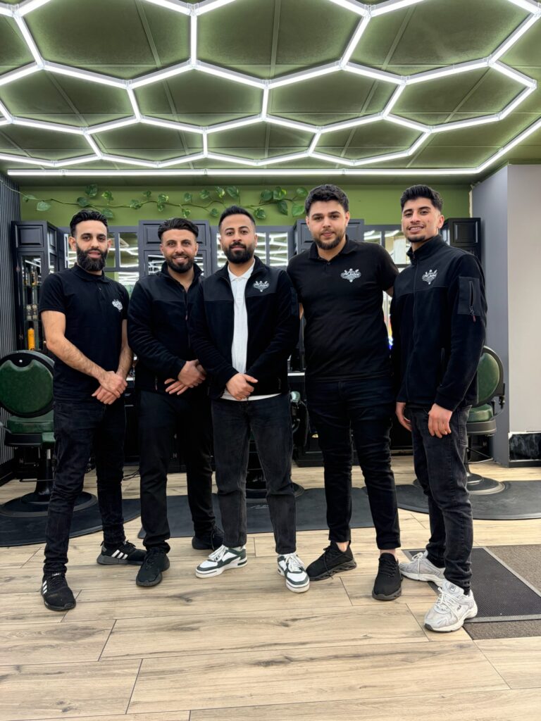 Fünf professionelle Barber in schwarzer Team-Kleidung stehen geschlossen und freundlich im modern beleuchteten Salon.
