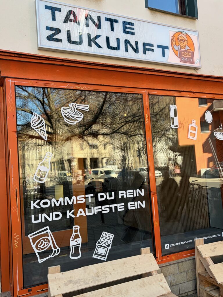 Außenansicht des 24/7-Ladens Tante Zukunft in Jena mit orangefarbener Fensterfront und dem Slogan „Kommst du rein und kaufste ein“.