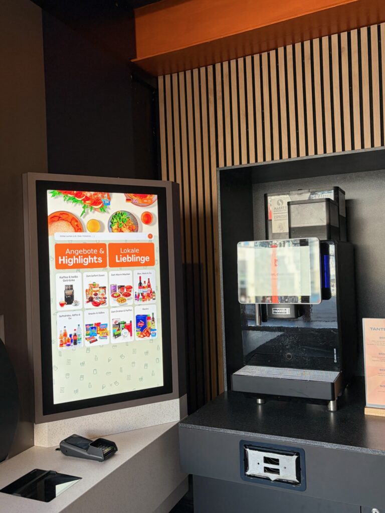 Ein großes digitales Touch-Terminal zur Auswahl von Produkten neben einer professionellen Kaffeemaschine im Innenraum von Tante Zukunft.