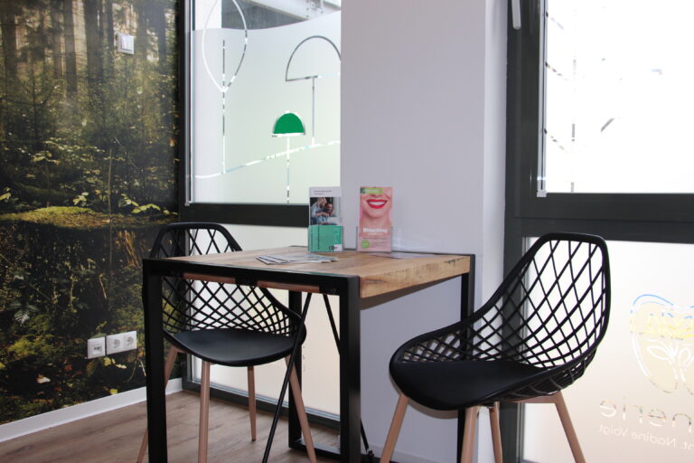 Moderner Wartebereich mit Holztisch, zwei schwarzen Stühlen und Informationsmaterial vor dem Fenster.