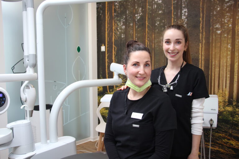 Zahnärztin Dr. Nadine Voigt und eine Mitarbeiterin vor der Behandlungseinheit und der Wald-Fototapete der Zähnerie.