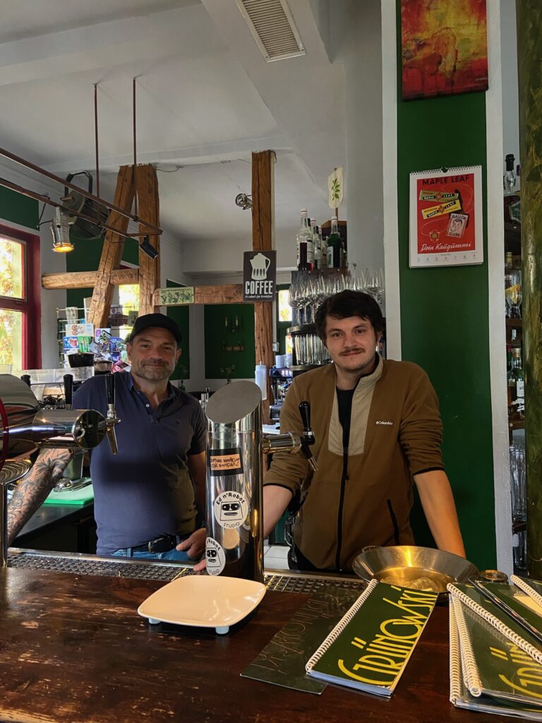 Inhaber stehen freundlich hinter dem Tresen, davor eine Zapfsäule mit Stickern; im Hintergrund grüne Wände und Regale.