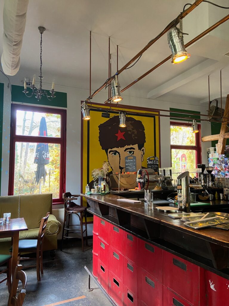 Blick auf den Barbereich mit roter Tresenverkleidung, großem gelben Wandbild und industrieller Beleuchtung an Kupferrohren.