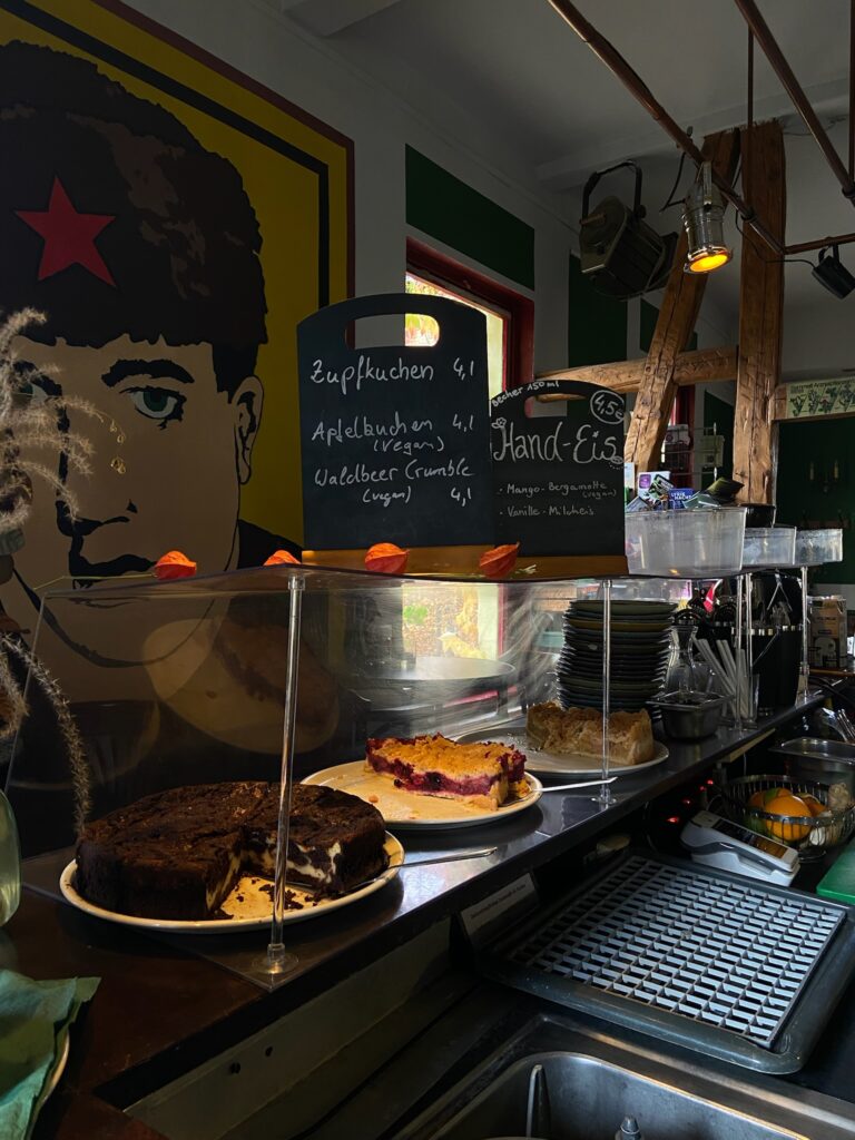 Kuchenvitrine mit handgeschriebenen Schildern für veganen Apfelkuchen und Zupfkuchen, im Hintergrund ein großes Pop-Art-Wandbild.