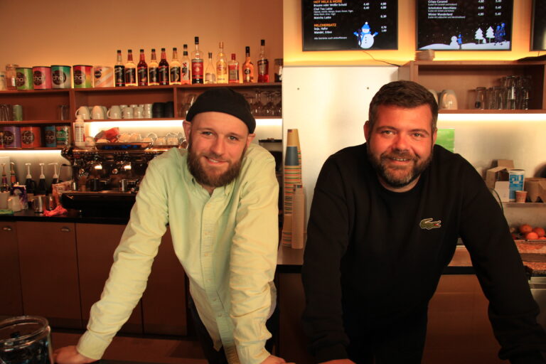 Inhaber stehen lächelnd hinter dem Tresen von The Bean, im Hintergrund die Espressomaschine und digitale Menüboards.