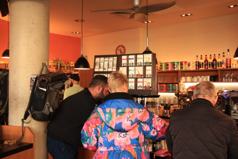 Blick in den belebten Gastraum von The Bean mit Kunden am Tresen, einer großen Auswahl an Althaus-Teesorten im Regal und Sirup-Flaschen.
