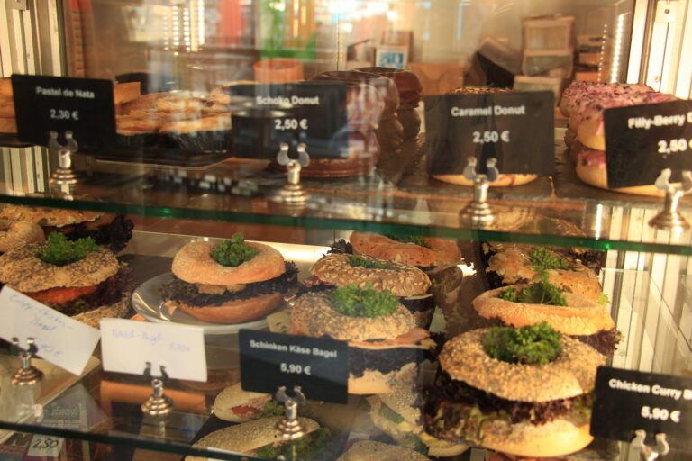 Glasvitrine gefüllt mit frisch belegten Bagels (z.B. Chicken Curry, Schinken-Käse), Donuts und Pastel de Nata.
