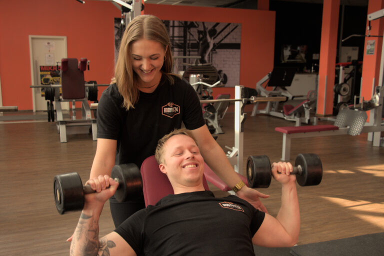 Eine Trainerin im Bodyfit-Shirt hilft lächelnd einem Mitglied beim Kurzhantel-Training auf der Schrägbank.