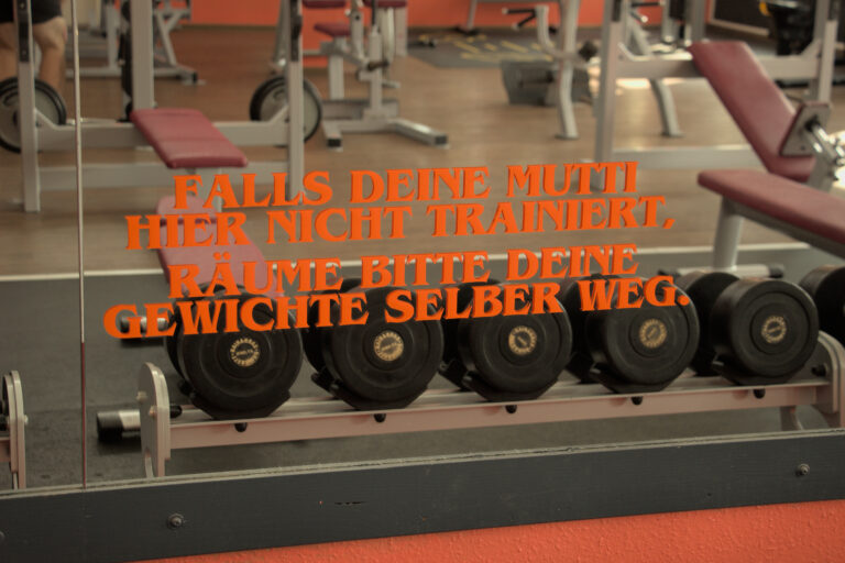 Kurzhantelablage vor einem Spiegel mit dem lustigen Spruch: "Falls deine Mutti hier nicht trainiert, räume bitte deine Gewichte selber weg."