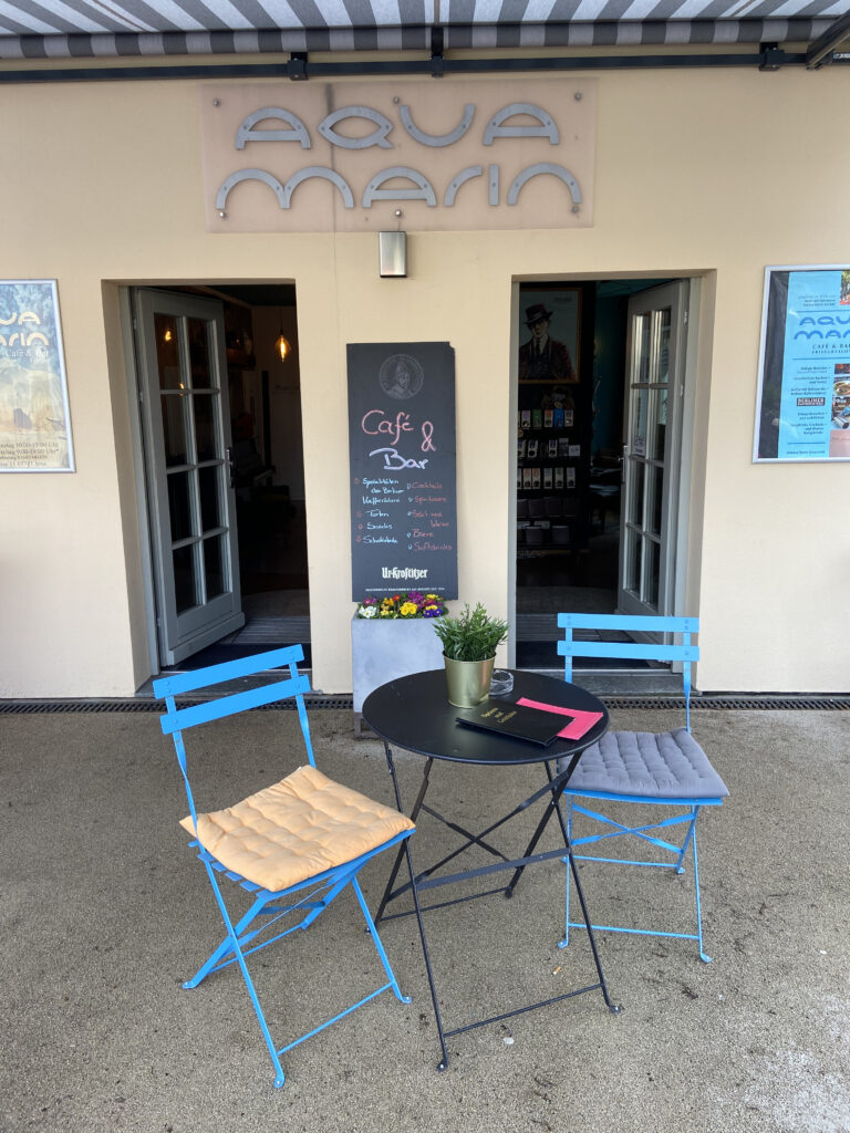 Eingang des Aquamarin Café & Bar in Jena mit beiger Fassade, großem Schriftzug über der Tür und blauen Bistrostühlen davor.