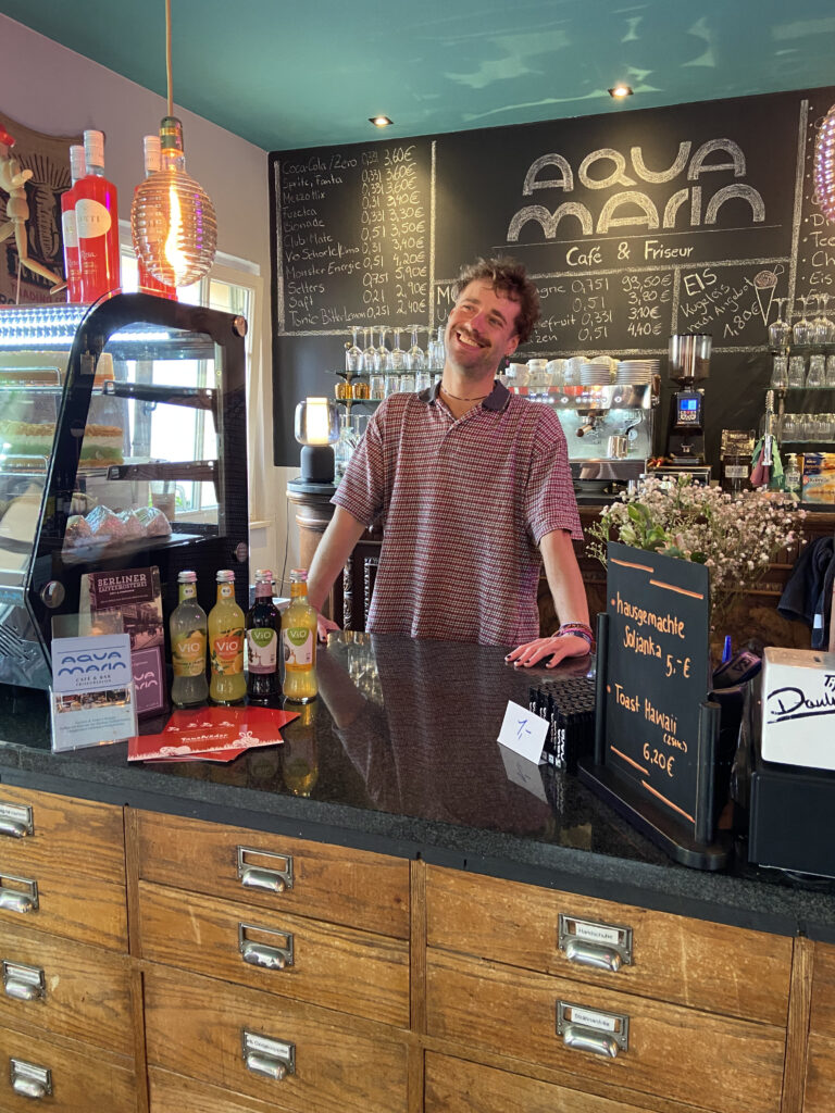 Freundlicher Mitarbeiter steht lächelnd hinter dem schwarzen Tresen im Aquamarin Café unter einer handgeschriebenen Angebotstafel.