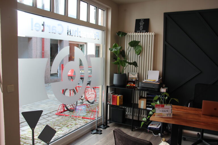 Innenraum der Fahrschule Cartel mit Schreibtisch, Pflanzen und großer Fensterfront mit Logo-Folie.